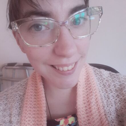 Photo of H.J., who has glasses on and is dressed in a floral top with a knitted cardigan and a peach coloured scarf around her neck. She has her hair up and is beaming at the camera.