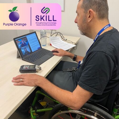 Photo of SKILL Project Leader, Andrew Gibson, sitting at a desk with a laptop open in front of him. He is sitting in a wheelchair and has his hands placed on the desk in front of him. In the top left corner are the Purple Orange and SKILL logos.