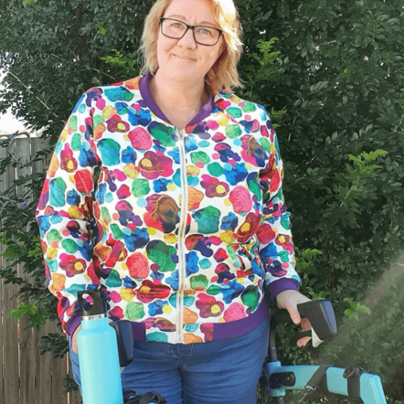 Photo of Michelle, a woman with white skin and a blonde bob, standing outdoors in front of a hedge. She wears a jacket covered in rainbow coloured paint splotches, denim jeans, and is holding onto a turquoise blue rollator. A handbag covered in rainbow splotches is attached to her rollator and in the cup holder to the side is a turquoise drink bottle.