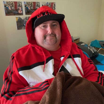 Photo of John at home. He is a man with white skin and facial stubble, and wears a black baseball cap with the AC/DC band logo, red shirt and a red hoodie, hood up over the cap.