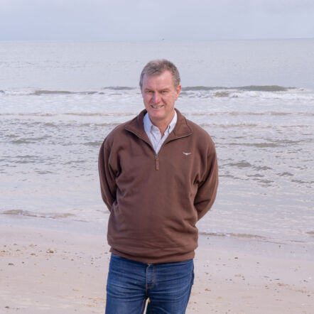 Photo of Gavin standing, with background of the sea and waves at the beach. He is a man with short grey hair, dressed in a brown jumper and jeans.