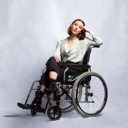 Studio photo of Jamila, who has white skin and brown hair in a short bob, and is dressed in a white flowy blouse and black pants. They sit confidently in their wheelchair, head resting on one hand.